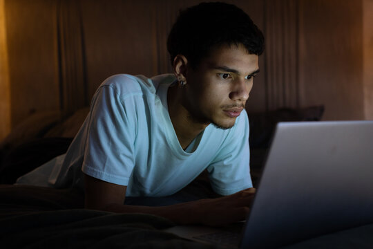 African American man lying on bed wearing white T-shirt using laptop in bedroom with lamp glow