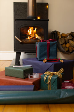 Stacked wrapped gifts are resting on floor near wood-burning stove, warm light casting reflections