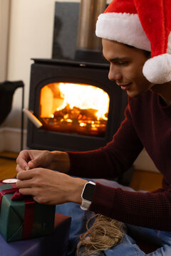 African American Santa-hatted man sitting on wooden floor by stove tying twine around wrapped gift