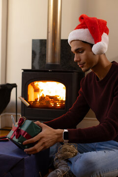 African American man wearing Santa hat sitting by wood stove wrapping gift with twine, copy space