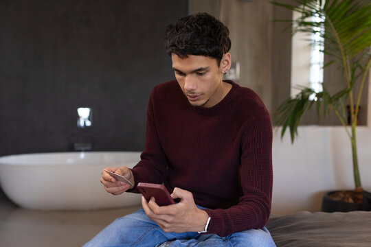 African American man sitting on cushioned bench in modern bathroom holding smartphone and bank card