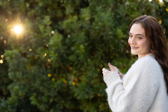 Woman turning toward camera in garden holding clear glass in white fuzzy sweater, copy space