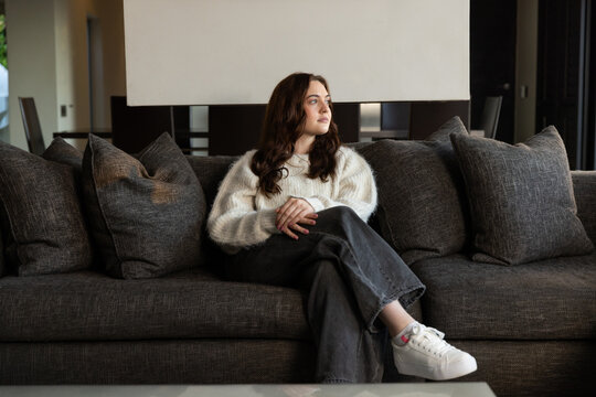 Female sitting on gray sofa in living room, wearing cream sweater and white sneakers