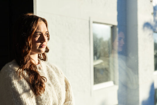 Woman in 20s standing by large glass window, sunlight reflecting on cream sweater, copy space