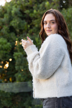 Woman standing on balcony holding glass cup by railing wearing fuzzy sweater and jeans, copy space