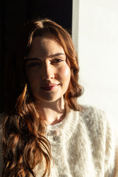 Woman sitting near bright window, sunlight splitting face, wearing cream sweater and pendant