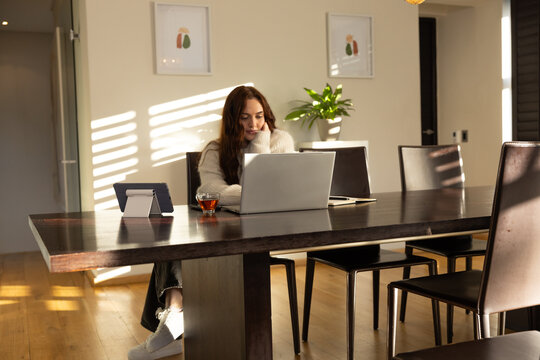 Adult woman wearing cozy sweater and jeans, sitting at dining table using laptop and tablet