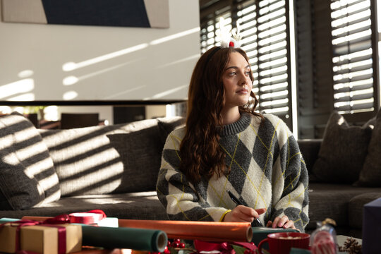 Adult woman sitting on gray sofa at home, wearing argyle sweater, writing gift tag among presents