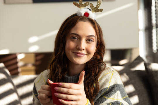 Adult woman sitting on sofa in living room holding red mug wearing knit sweater glitter antlers