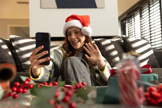 Woman sitting on couch at home wearing Santa hat and knit sweater, holding smartphone, waving