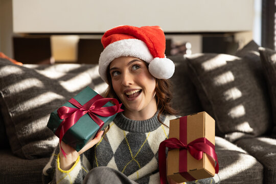 Woman sitting on sofa at home wearing Santa hat, argyle sweater holding two red-ribboned gifts