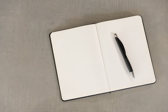 Open notebook lying on textured grey tabletop with blank pages and black pen resting diagonally