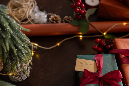 Green wrapped gift is resting on dark wooden table, showing kraft tag and warm string lights
