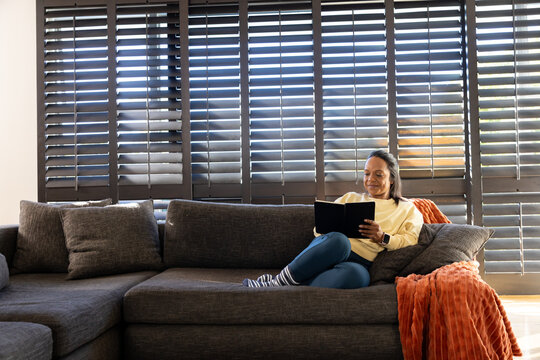 Mature African woman lounging on gray sofa holding black tablet, reading by shutters, copy space