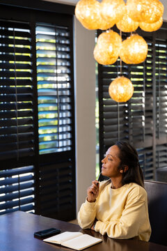 Mature African woman sitting at table with pen looking through shutters notebook phone, copy space