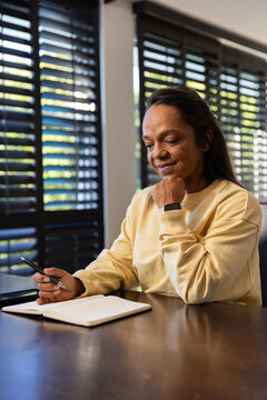 African American woman in yellow sweatshirt sitting at table at home holding pen over notebook