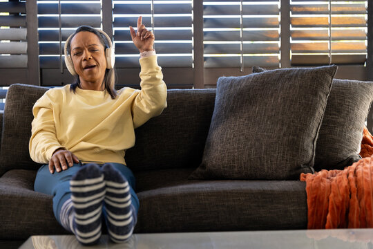 Mature African American woman sitting, listening and singing on sofa with headphones, striped socks