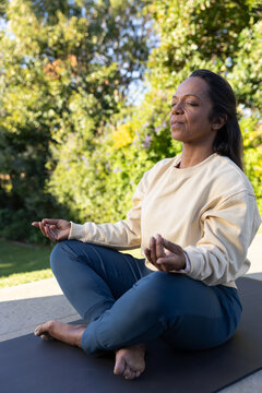Mature African American woman meditating on patio yoga mat, wearing cream top and blue leggings