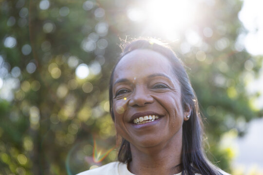 Mature African woman smiling, facing camera in garden wearing light top, stud earrings, lens flare