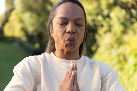 African American woman standing in sunlit garden, joining palms in prayer, wearing cream sweatshirt