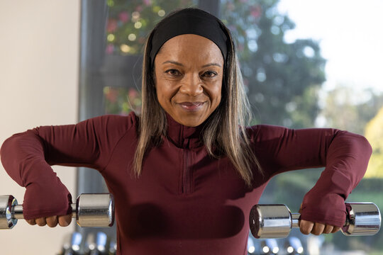 African American woman holding chrome dumbbells exercising in maroon top in home gym by window