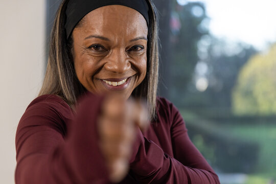 Mature African woman smiling extending arm toward lens by window in black headband and maroon top