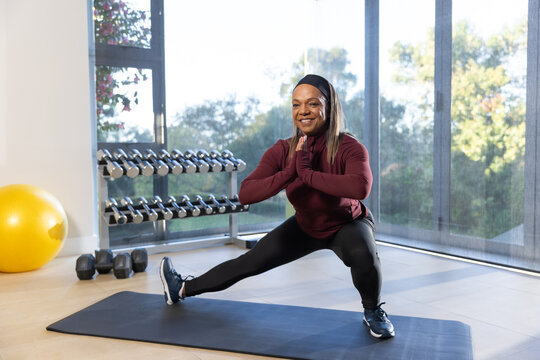 Mature African American woman in maroon top doing lateral lunge in home gym with dumbbells, mat