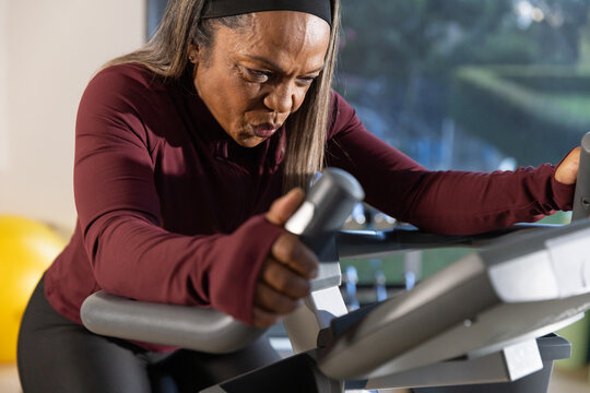 Mature woman in maroon top black leggings gripping bars pedaling on spin bike at gym console