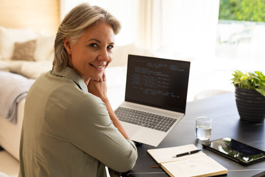 Senior woman smiling, typing on laptop at home workspace with notebook, pen, tablet, plant, sofa