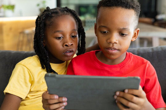 African American children sitting on dark gray sofa in living room, leaning in, watching tablet