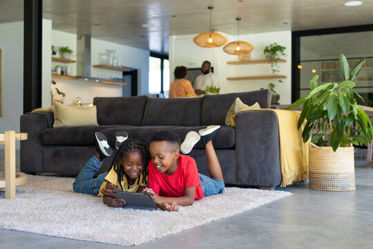 African American children lying on rug at home sharing tablet, wearing red shirt and yellow top