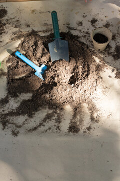 Potting soil mound sitting on light sheet with green trowel, blue mini-rake, peat pot part-filled