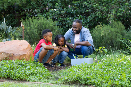 African American family crouching tending plants in yard with white planter and green spray bottle