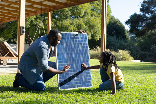 African American man and child kneeling on grassy backyard lawn, examining photovoltaic solar panel