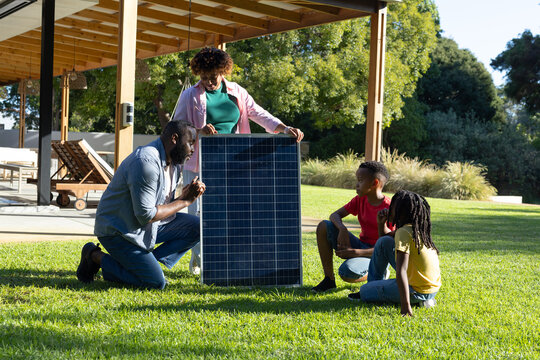 African American family kneeling, standing, sitting, installing solar panel on lawn near pergola