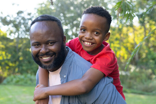 African American father and son smiling, sharing piggyback in sunlit grassy park, light blue shirt