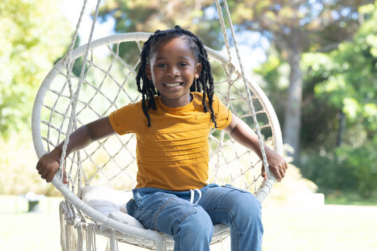 African American girl sitting in macrame swing holding ropes, wearing mustard top and jeans in yard