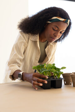 Adult woman leaning over table, examining seed tray, peat pots, wearing beige shirt and smartwatch