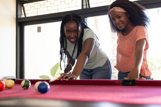 African female friends leaning over magenta pool table at home, taking shot with cue and balls