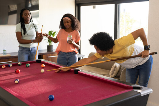 African American female friends playing pool at home near red felt table with billiard balls, jar