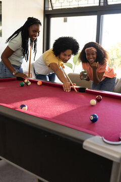 African American friends playing pool in recreation room, man lining up shot with wooden cue stick