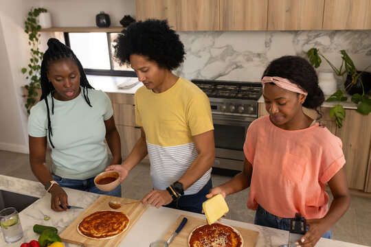 Three friends preparing pizzas at kitchen island with wooden boards, sauce bowl, grater, vegetables