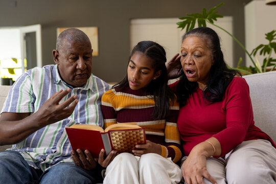 African American grandparents with teenage granddaughter sitting on sofa at home holding red book