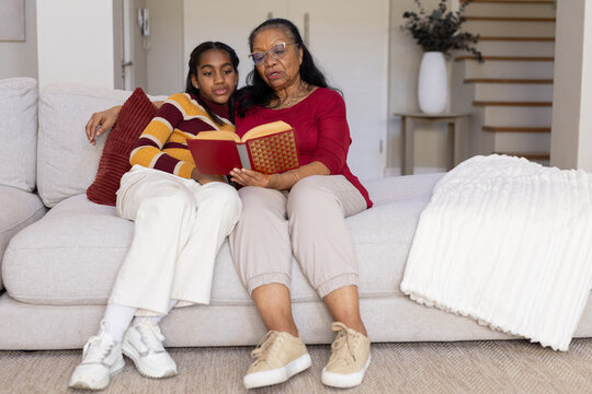 Senior woman and teen girl sitting on light gray sofa in living room reading red book