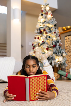 African American teen lying on carpet at home reading red book, patterned socks near Christmas tree