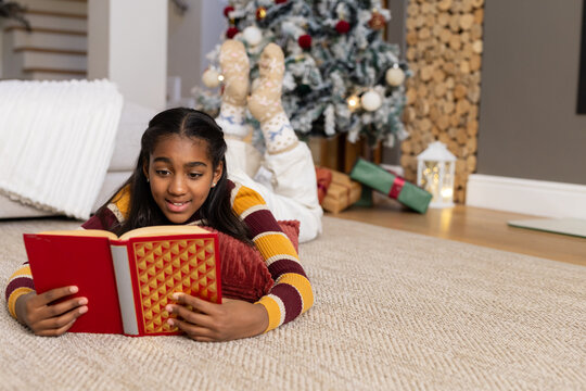 African American teen female lying on rug at home reading red hardback book near Christmas tree