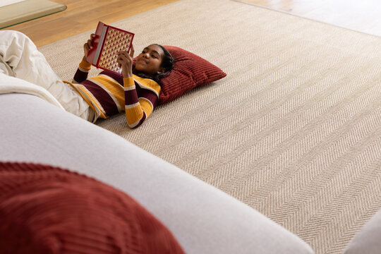 Youth African American in striped sweater lying on herringbone rug in living room holding red book