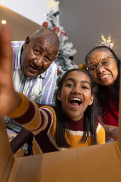 Grandparents leaning while youth granddaughter reaching into open cardboard box at home, tree lit
