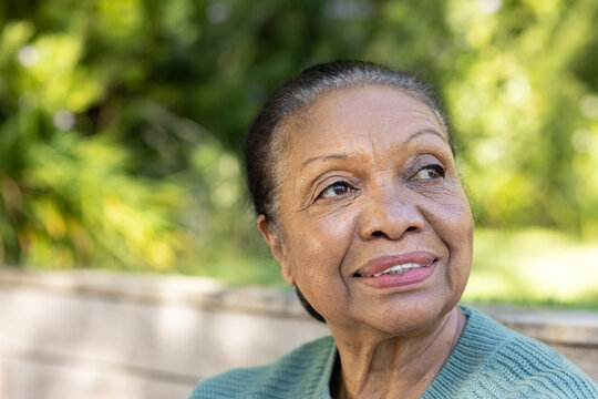 Senior African American woman sitting on wooden bench in leafy park, wearing green knit sweater