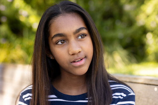 African American female teen sitting on wooden bench in garden, wearing navy striped top, studs